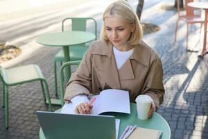 Image of young woman studying outdoors, sitting in cafe, making notes while listening to online lecture using laptop, making notes in notebook, writing photo