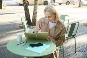 Portrait of beautiful blond woman, sitting in outdoor coffee shop, drawing in cafe in notebook, making sketches outside on street photo