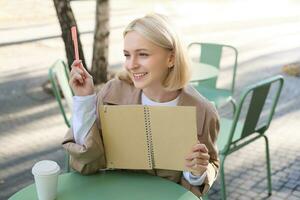 Portrait of young beautiful blond woman, artsy girl in coffee shop, holding notebook and pen, writing in her journal, drawing sketches outdoors photo