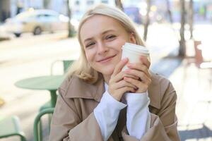 Close up portrait of smiling blond woman, holding cup of coffee, sitting in outdoor, street cafe, looking happy, waiting for someone in restaurant outside photo