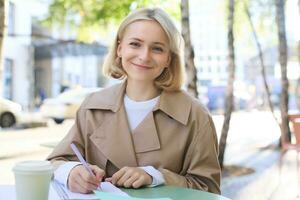 Portrait of young smiling woman, student working, doing homework outdoors, sitting in street cafe, drinking coffee from takeaway cup photo