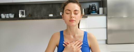 Image of calm and relaxed woman meditating, doing breathing practices, holding hands on chest during yoga session at home photo