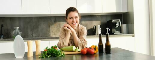 Portrait of smiling young woman cooking in kitchen in cosy clothes, standing near counter with chopping board, vegetables, zucchini, preparing dinner for family, making salad photo