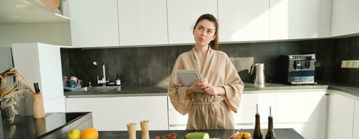 retrato de creativo joven mujer, cocinando, participación receta libro, pensando mientras haciendo comida, preparando comida en el cocina, en pie en bata de baño foto