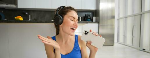 Close up portrait of young happy woman, workout at home, listens music in wireless headphones, singing at smartphone, doing training exercises photo