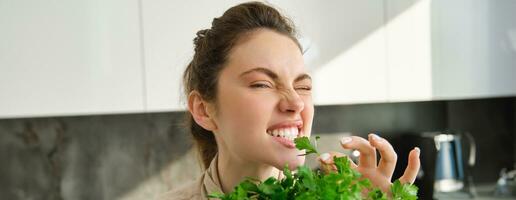 Funny and cute brunette woman bites delicious, fresh green parsley, eating herbs, cooking salad in kitchen photo