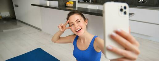 cerca arriba retrato de hermosa morena deportista, tomando selfie en teléfono inteligente, posando en aptitud estera con móvil teléfono, registros su aptitud ejercicios, rutina de ejercicio formación foto