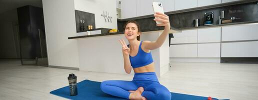 activo joven mujer, vlogger lo hace Deportes, registros su rutina de ejercicio formación desde hogar en teléfono inteligente cámara, posando para selfie dentro su casa, se sienta en caucho yoga estera en azul polainas y sujetador deportivo foto