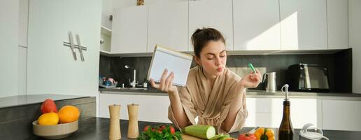 Portrait of modern young woman cooking, making grocery list, reading recipe and making meal, salad in the kitchen, looking at vegetables on chopping board photo