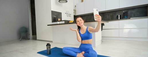 Active young woman does sports, records her workout training from home on smartphone camera, posing for selfie inside her house, sits on rubber yoga mat in blue leggings and sportsbra photo