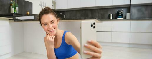 Woman doing workout training from home and taking selfie on smartphone camera, posing for photo inside her house, sits on rubber yoga mat in blue leggings and sportsbra