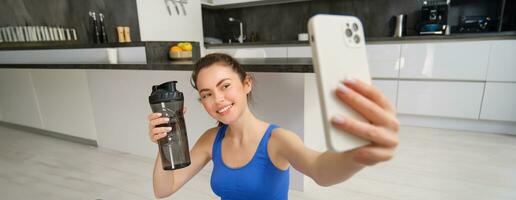 Portrait of sportswoman takes selfie with water bottle in living room, holds smartphone and poses for photo while doing fitness workout