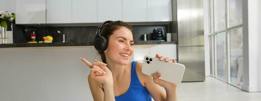 Close up portrait of young happy woman, workout at home, listens music in wireless headphones, singing at smartphone, doing training exercises photo