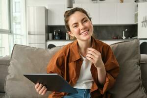 retrato de joven morena mujer leyendo en digital tableta, acecho televisión serie en su solicitud, sentado en sofá en vivo habitación foto