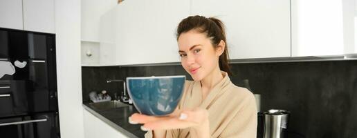 Portrait of beautiful, smiling young woman offering you morning cup of coffee, extending her hand with mug to you, standing in bathrobe in the kitchen photo