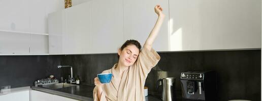 retrato de contento niña bailando con café en el cocina, vistiendo bata de baño, disfrutando su Mañana rutina foto