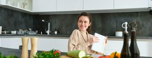 Portrait of woman writing down list of groceries, making notes in recipe, sitting in kitchen near vegetables, preparing dinner menu photo