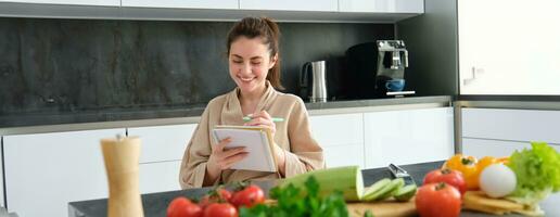 Portrait of beautiful, smiling young woman making list of meals, writing down recipe, sitting in the kitchen with vegetables, doing house errands photo
