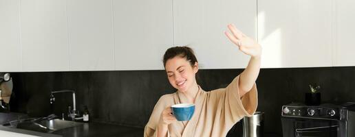 Portrait of beautiful, happy young woman covers her eyes from morning sunlight, waking up and drinking coffee, holding mug, posing in the kitchen photo
