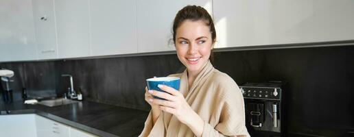 estilo de vida concepto. retrato de contento morena mujer en bata de baño, Bebiendo café en el cocina, teniendo Mañana taza y sonriente foto