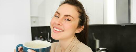 Portrait of happy, smiling modern woman, starts her day with morning mug of tea. Girl drinking coffee in the kitchen, standing with mug and looking relaxed photo