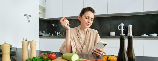 Image of young beautiful woman, holding tomato, sitting in kitchen with smartphone, chopping board and vegetables on counter, cooking food, order groceries for her recipe, using mobile phone app photo