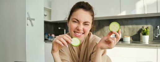 Portrait of beautiful brunette girl cooking in the kitchen, posing in bathrobe at home, holding zucchini, showing happy smile, making healthy food, vegetarian meal photo