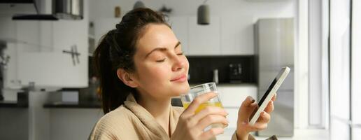 Close up of beautiful smiling woman, drinks fresh orange juice, enjoys her morning, holds mobile phone photo