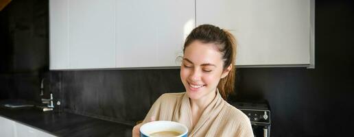 Lifestyle concept. Portrait of happy brunette woman in bathrobe, drinking coffee in the kitchen, having morning cuppa and smiling photo