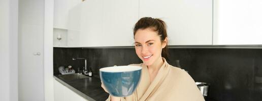 retrato de hermoso, sonriente joven mujer ofrecimiento usted Mañana taza de café, extensión su mano con jarra a tú, en pie en bata de baño en el cocina foto