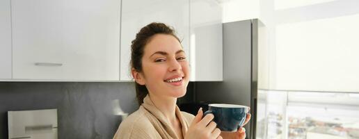 retrato de contento joven mujer empieza su Mañana con jarra de café, Bebiendo té desde taza, en pie en el cocina, sonriente alegremente foto