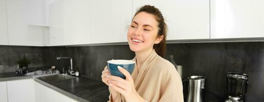 retrato de contento joven mujer empieza su Mañana con jarra de café, Bebiendo té desde taza, en pie en el cocina, sonriente alegremente foto