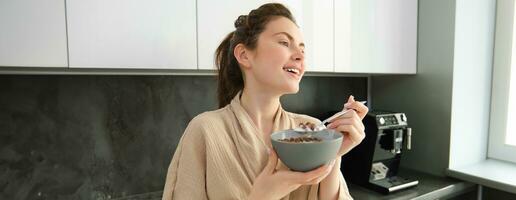 Good-looking brunette woman eating her breakfast, standing in kitchen near worktop and holding bowl of cereals with milk, enjoying her morning, wearing cosy bathrobe photo
