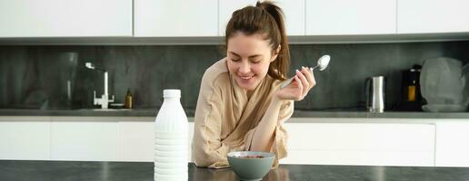 retrato de contento joven mujer se inclina en cocina encimera y comiendo cereales, tiene Leche y cuenco en frente de su, teniendo su desayuno, vistiendo bata de baño foto