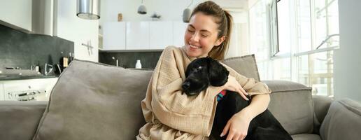 Happiness and lifestyle concept. Adorable dog spends time with its owner, young woman hugging her puppy on sofa at home photo