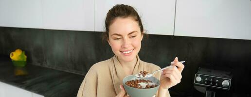 Happy mornings. Gorgeous young woman eating cereals with milk, standing in kitchen with breakfast bowl, enjoying start of the day, smiling photo