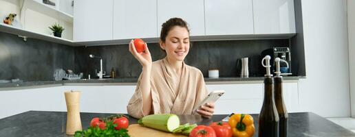 Image of young beautiful woman, holding tomato, sitting in kitchen with smartphone, chopping board and vegetables on counter, cooking food, order groceries for her recipe, using mobile phone app photo