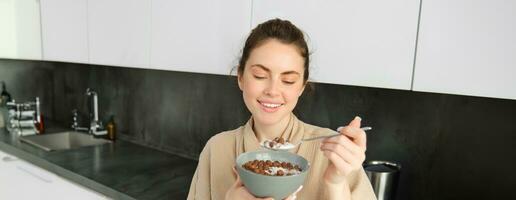 Close up portrait of smiling, beautiful woman eating cereals with milk, holding spoon and bowl, looking outside window in morning, standing near kitchen worktop photo