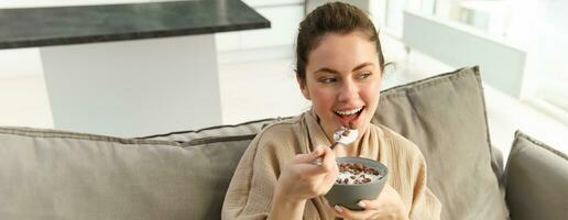 Portrait of woman on sofa in bathrobe, eating cereals with milk, watching tv and having breakfast photo