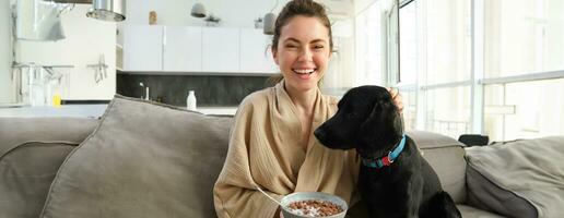 Happy mornings. Beautiful woman enjoying her morning, sitting on sofa, eating breakfast and playing, cuddling with dog, puppy asks for small bite of cereals photo