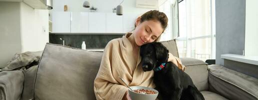 woman with short hair sitting on a couch at home, eating cereals for breakfast, and petting her beloved dog. The dog is sniffing a bowl of cereals photo