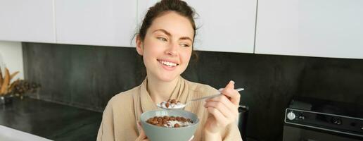 Happy mornings. Gorgeous young woman eating cereals with milk, standing in kitchen with breakfast bowl, enjoying start of the day, smiling photo