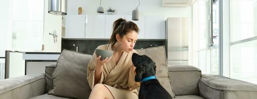 Happy young woman in bathrobe, enjoys spending time with her dog at home, eating cereals, having breakfast with puppy, sitting on sofa photo