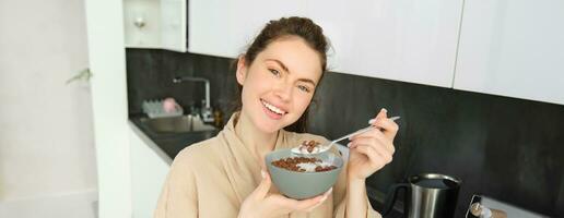 Close up of beautiful smiling woman in bathrobe, standing in kitchen near worktop, eating bowl of chocolate cereals with milk, holding spoon and looking happy at camera photo