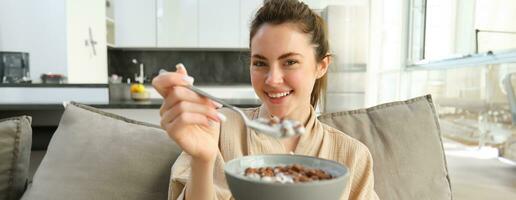 Image of smiling, happy young woman eating breakfast, holding bowl of cereals with milk, having meal at home photo