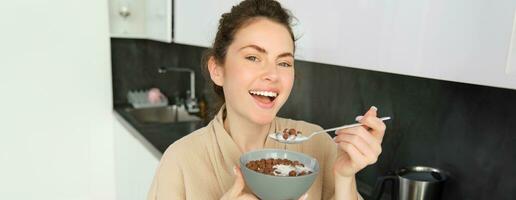 Happy mornings. Gorgeous young woman eating cereals with milk, standing in kitchen with breakfast bowl, enjoying start of the day, smiling photo