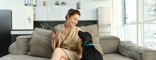 Home and lifestyle concept. Beautiful young woman with dog, sitting on sofa, eating cereals and hugging her puppy, girl having breakfast in bathrobe photo