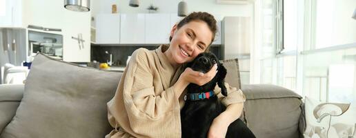 Young woman hugging adorable dog, cuddling her puppy on sofa at home photo