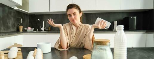 Attractive young cheerful girl baking at the kitchen, making dough, holding recipe book, having ideas photo