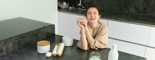 Attractive young cheerful girl baking at the kitchen, making dough, holding recipe book, having ideas photo